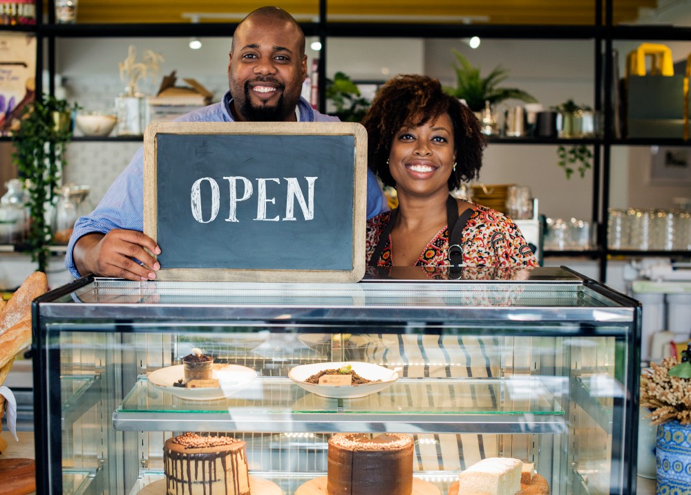 A bakery couple holding an OPEN sign at their counter